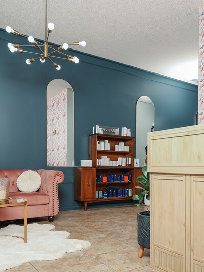 picture of clinic with desk off to right and pink couch on left with brown cabinet full of skin care products.  Wall behind skin care is navy blue with mirrors